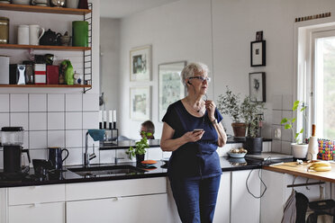 Retired senior woman holding smart phone while listening to in-ear headphones in kitchen at home - MASF07411