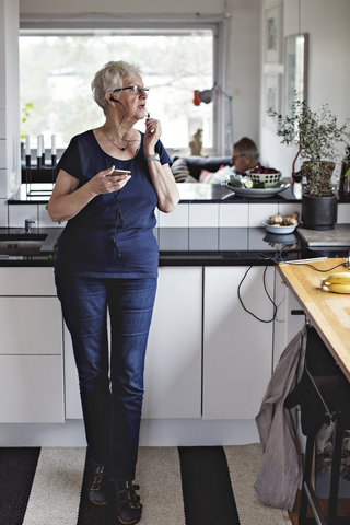 Ältere Frau in voller Länge stehend mit Smartphone in der Küche zu Hause, lizenzfreies Stockfoto