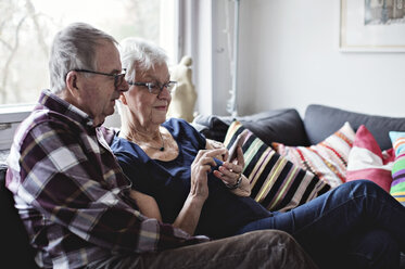 Älteres Paar im Ruhestand, das sich ein Smartphone teilt und auf dem Sofa im Wohnzimmer sitzt - MASF07413