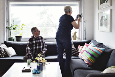 Älterer Mann betrachtet Frau, die eine Stehlampe am Sofa im Wohnzimmer einstellt - MASF07415
