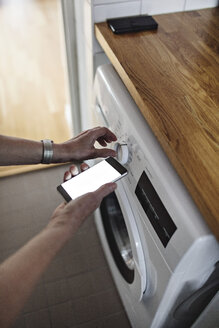 Cropped hands of senior woman holding mobile phone while using washing machine in bathroom at home - MASF07417