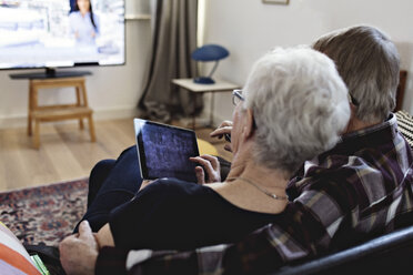 Rear view of senior couple sitting with digital tablet watching TV in living room at home - MASF07431