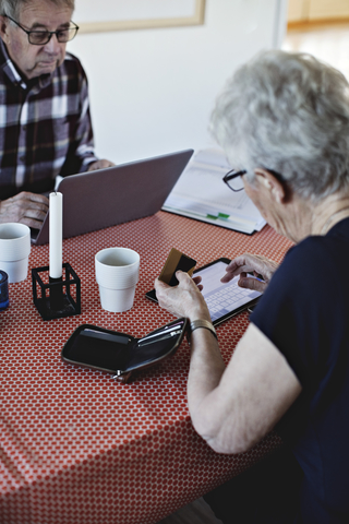 Ältere Frau hält eine Kreditkarte, während sie am Esstisch ein digitales Tablet benutzt, lizenzfreies Stockfoto