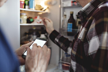 Senior woman holding smart phone standing by retired man in kitchen at home - MASF07443