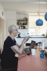 Seitenansicht einer älteren Frau im Ruhestand, die ein Smartphone benutzt, während sie mit einem Laptop am Esstisch sitzt - MASF07449