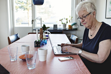 Ältere Frau, die einen Laptop benutzt, während sie am Esstisch im Zimmer sitzt - MASF07456