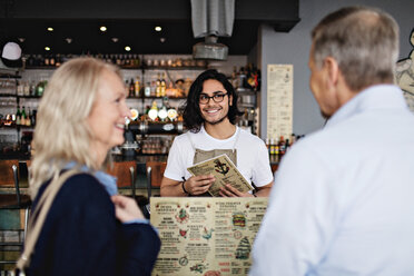 Lächelnder Besitzer schaut auf reifes Paar mit Speisekarte im Restaurant - MASF07496