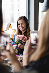 Freundinnen fotografieren lächelnde Frau mit Blumenstrauß am Esstisch im Restaurant - MASF07498