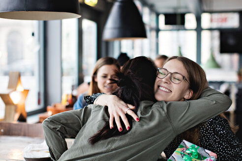 Lächelnde Freundinnen, die sich umarmen und ein Geburtstagsgeschenk halten, während sie im Restaurant sitzen - MASF07520