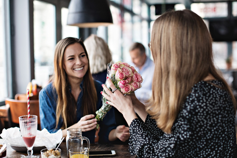 Lächelnde junge Frau schenkt einer Freundin im Restaurant einen frischen Blumenstrauß, lizenzfreies Stockfoto