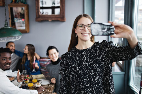Lächelnde junge Frau nimmt Selfie mit Freunden sitzen im Restaurant - MASF07542