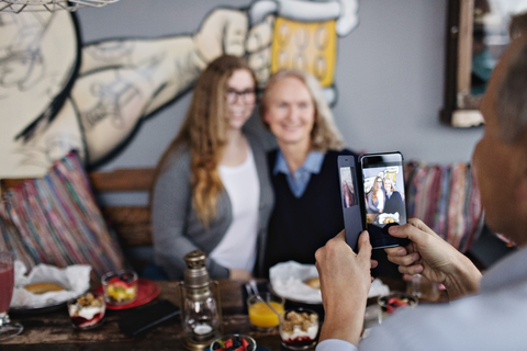 Älterer Mann fotografiert Frauen mit seinem Smartphone im Restaurant, lizenzfreies Stockfoto