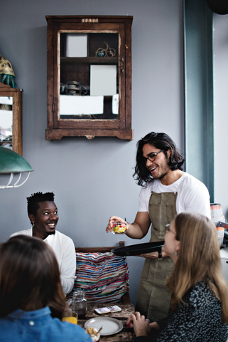 Ein lächelnder Besitzer serviert einem jungen Mann und einer Frau am Esstisch das Essen, lizenzfreies Stockfoto
