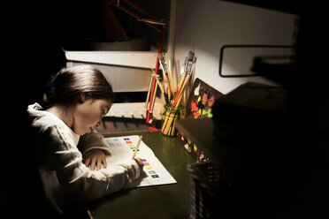 Blick von oben auf ein Mädchen, das an einem beleuchteten Schreibtisch in einer Dunkelkammer sitzt und lernt - MASF07580