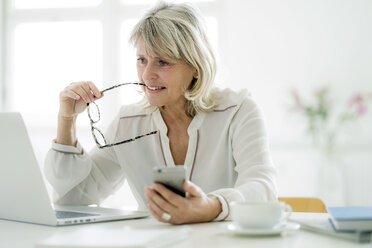 Mature businesswoman holding cell phone working on laptop at desk - HHLMF00250