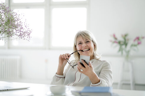 Porträt einer glücklichen reifen Geschäftsfrau, die ein Mobiltelefon am Schreibtisch hält - HHLMF00252