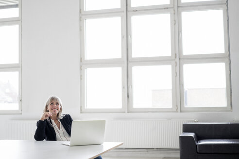 Lächelnde reife Geschäftsfrau mit Laptop am Schreibtisch im Büro und Blick nach oben - HHLMF00267