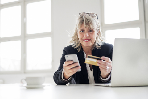 Mature businesswoman with smartphone, credit card and laptop at desk in the office stock photo