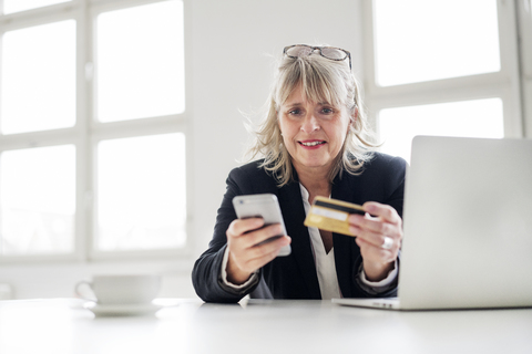 Reife Geschäftsfrau mit Smartphone, Kreditkarte und Laptop am Schreibtisch im Büro, lizenzfreies Stockfoto