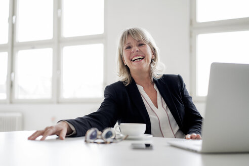 Portrait of laughing mature businesswoman with laptop at desk in the office - HHLMF00273