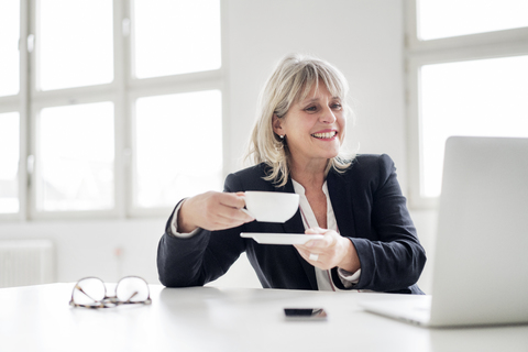 Lächelnde reife Geschäftsfrau mit einer Tasse Kaffee, die an einem Laptop am Schreibtisch im Büro arbeitet, lizenzfreies Stockfoto