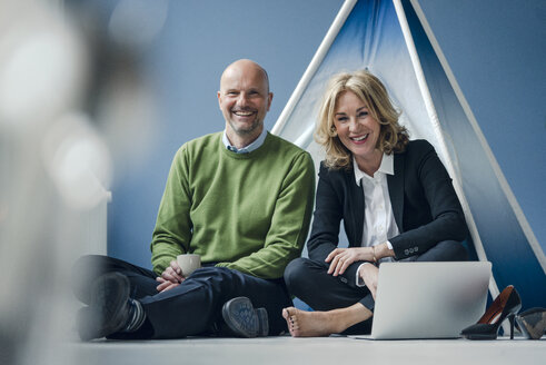 Happy businessman and businesswoman sitting at teepee indoors with laptop - KNSF03818