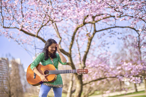 Glückliche junge Frau mit Gitarre in einem Park an einem Kirschblütenbaum, lizenzfreies Stockfoto
