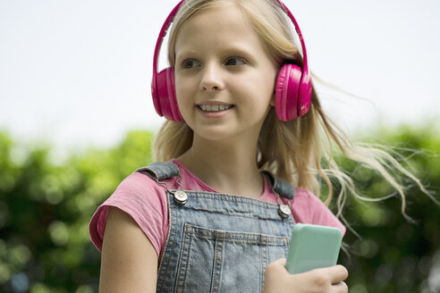 Nettes blondes Mädchen hört Musik mit rosa Kopfhörern im Garten - SBOF01479