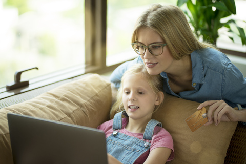 Mutter und Tochter im modernen Wohnzimmer auf einer Couch mit Laptop und Kreditkarte, lizenzfreies Stockfoto