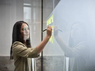 Businesswoman writing on sticky notes at glass pane in office - CVF00370