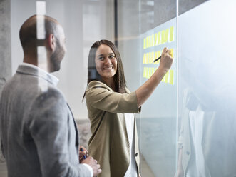 Businesswoman smiling at colleague writing on sticky notes at glass pane in office - CVF00371