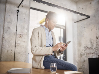 Businessman sitting on desk in office with cell phone and earbuds - CVF00386