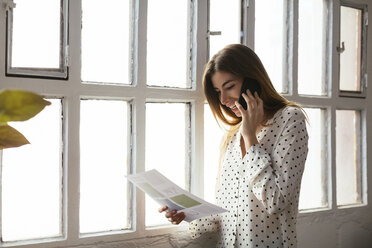 Glückliche junge Frau mit Handy, die ein Dokument am Fenster betrachtet - EBSF02495