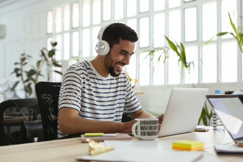 Lachender junger Mann mit Kopfhörern und Laptop am Schreibtisch im Büro, lizenzfreies Stockfoto