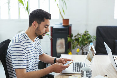 Junger Mann mit Laptop und Mobiltelefon am Schreibtisch im Büro - EBSF02511