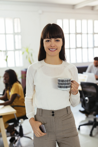 Porträt einer lächelnden jungen Frau im Büro mit Kollegen im Hintergrund, lizenzfreies Stockfoto