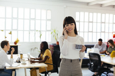 Portrait of smiling young woman on cell phone in office with colleagues in background - EBSF02527