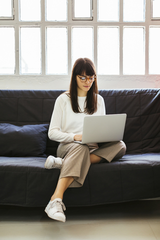 Junge Frau sitzt auf einer Couch im Büro und benutzt einen Laptop, lizenzfreies Stockfoto