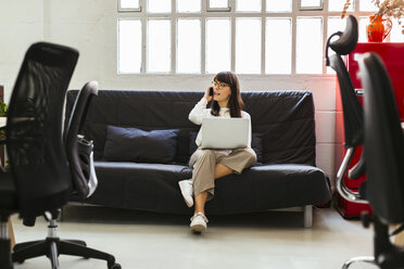Junge Frau sitzt auf der Couch im Büro und benutzt Handy und Laptop - EBSF02543