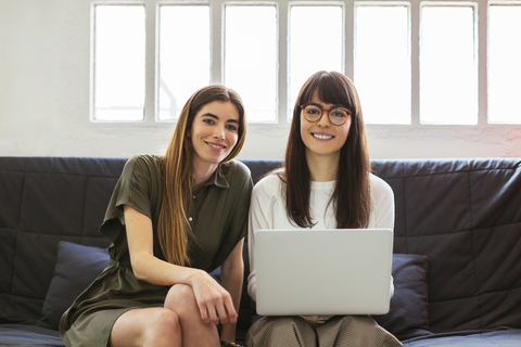 Zwei lächelnde junge Frauen sitzen auf einer Couch im Büro mit Laptop, lizenzfreies Stockfoto