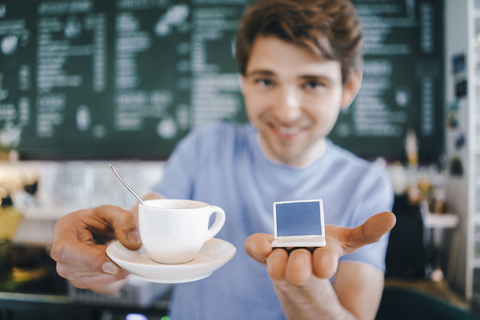 Lächelnder Mann in einem Café bietet eine Tasse Kaffee an und hält ein Miniatur-Laptop-Modell, lizenzfreies Stockfoto