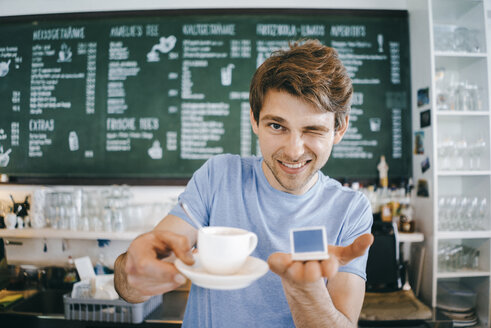 Lächelnder Mann in einem Café bietet eine Tasse Kaffee an und hält ein Miniatur-Laptop-Modell - KNSF03893