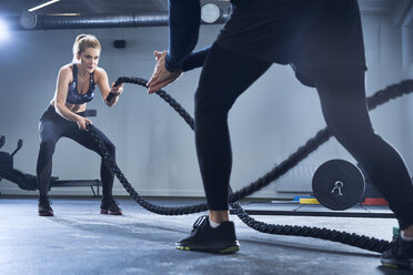 Athletic woman doing battlerope exercise with personal trainer at gym - BSZF00356