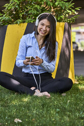 Portrait of smiling woman sitting barefoot on a meadow listening music with headphones and cell phone - BEF00034