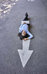 Laughing young businesswoman with laptop lying on arrow sign on street - BEF00037