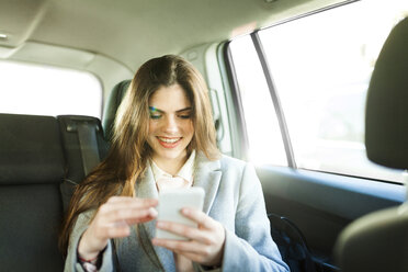 Porträt einer lächelnden jungen Geschäftsfrau, die auf dem Rücksitz eines Autos sitzt und auf ihr Handy schaut - VABF01568