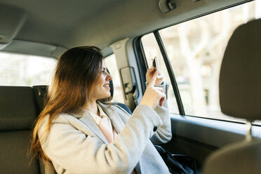 Smiling young woman sitting on backseat of a car taking picture with cell phone - VABF01575