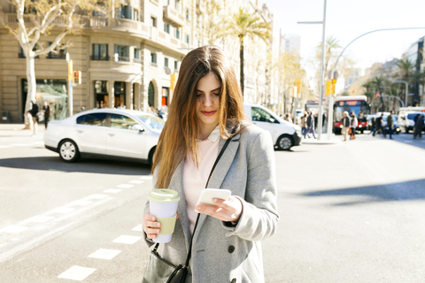 Spanien, Barcelona, junge Frau mit Kaffee zum Mitnehmen steht am Straßenrand und schaut auf ihr Handy, lizenzfreies Stockfoto