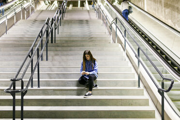 Junge Frau sitzt auf einer Treppe und benutzt Handy und Kopfhörer - VABF01609