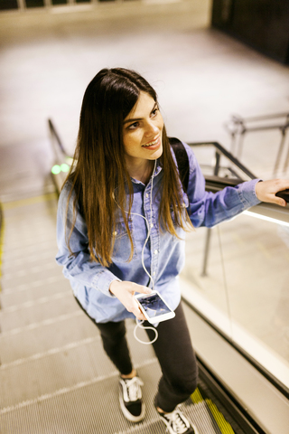 Porträt einer lächelnden jungen Frau, die auf einer Rolltreppe steht und mit einem Mobiltelefon und Kopfhörern Musik hört, lizenzfreies Stockfoto
